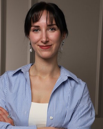 Junge Frau mit langen, dunklen Haaren und Ohrringen, trägt ein gestreiftes Hemd und steht mit verschränkten Armen.