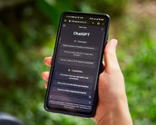 Ein Smartphone wird in einer Hand gehalten, auf dem Bildschirm ist die ChatGPT-Oberfläche mit Beispielen und Fähigkeiten sichtbar. Der Hintergrund zeigt unscharfe grüne Pflanzen.