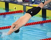 Ein Schwimmer springt kopfüber ins Wasser eines Schwimmbeckens, während er sich auf einen Wettkampf vorbereitet. Rote Schwimmbahnmarkierungen sind sichtbar.