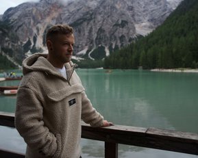 Ein Mann in einem beigen Fleece-Pullover steht an einem Holzgeländer und blickt auf einen grünen See, umgeben von Bergen und Wäldern.