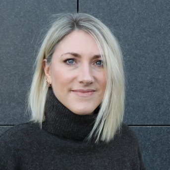 Eine Frau mit blonden, schulterlangen Haaren und einem dunklen Rollkragenpullover steht vor einer grauen Wand.