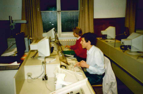 Zwei Personen sitzen an Computern in einem Büro, während sie an ihren Bildschirmen arbeiten.