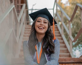 Eine junge Frau mit einem Abschlusshut sitzt auf einer Treppe und lächelt. Sie trägt eine akademische Robe und mehrere Medaillen um den Hals.