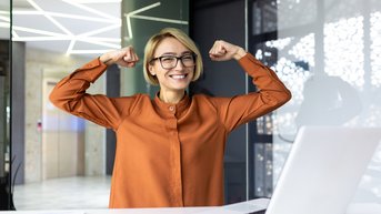 Eine lächelnde Frau mit Brille in einem modernen Büro hebt die Arme in einer Siegerpose. Vor ihr steht ein Laptop auf dem Tisch, daneben liegen ein Notizbuch und Stifte.