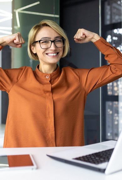Eine lächelnde Frau mit Brille in einem modernen Büro hebt die Arme in einer Siegerpose. Vor ihr steht ein Laptop auf dem Tisch, daneben liegen ein Notizbuch und Stifte.