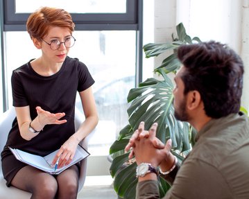Klinische Psychologie und psychische Erkrankungen Eine Frau mit Brille sitzt auf einem Stuhl und spricht mit einem Mann, der ihr gegenüber sitzt. Beide scheinen in ein Gespräch vertieft zu sein, umgeben von Pflanzen und natürlichem Licht.