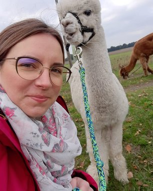 Eine Frau mit Brille posiert neben einem weißen Alpaka, das an einer Leine gehalten wird. Im Hintergrund ist ein weiteres Alpaka zu sehen. Die Frau trägt einen bunten Schal und eine rote Jacke.