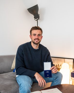 Ein Mann sitzt auf einem Sofa und hält eine Schmuckschatulle in der Hand. Er trägt einen blauen Pullover und hat einen neutralen Gesichtsausdruck. Im Hintergrund sind Bücherregale und eine Lampe sichtbar.
