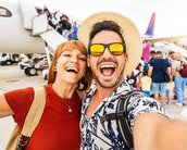 Ein Mann und eine Frau machen ein Selfie am Flughafen, während Passagiere im Hintergrund ein Flugzeug betreten. Beide lächeln und tragen Sonnenbrillen, der Mann hat einen Strohhut auf.
