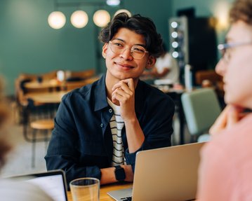 Ein junger Mann mit Brille sitzt an einem Tisch in einem Café, lächelt und schaut nachdenklich, während er mit anderen spricht.