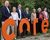 Eine Gruppe von sieben Personen steht nebeneinander und hält große orangefarbene Buchstaben, die das Wort "care" bilden. Sie tragen formelle Kleidung und stehen in einer grünen Umgebung.
