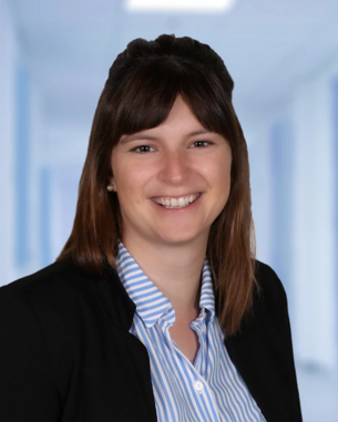 Eine lächelnde Frau mit langen, braunen Haaren trägt ein schwarzes Blazer und ein gestreiftes Hemd. Der Hintergrund ist unscharf und zeigt einen hellen, modernen Raum.