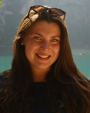 Eine Frau mit langen, braunen Haaren und Sonnenbrille lächelt vor einem klaren, türkisfarbenen See und bewaldeten Bergen.