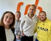 Vier Personen posieren gemeinsam vor einer Wand mit einem orangefarbenen Logo. Sie lächeln und tragen unterschiedliche Kleidung, darunter ein gelbes Sweatshirt.