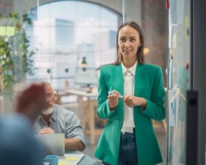 Eine Frau in einem grünen Blazer steht vor einer Gruppe, hält einen Stift und spricht. Im Hintergrund sind Notizen und ein Büro sichtbar.