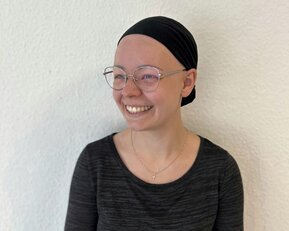 Eine lächelnde Person mit einer schwarzen Kopfbedeckung und Brille, die ein schwarzes Oberteil trägt, steht vor einer weißen Wand.