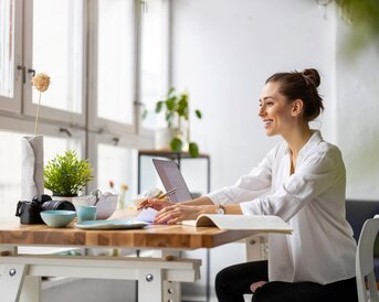 Eine Frau sitzt an einem Tisch mit einem Laptop, Notizbuch und Pflanzen. Sie lächelt und schreibt, während Tageslicht durch große Fenster strömt.