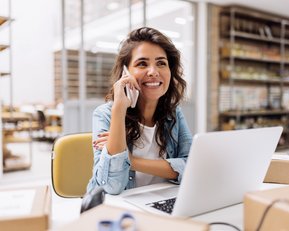 Eine Frau mit lockigem Haar sitzt an einem Schreibtisch, spricht am Telefon und lächelt. Vor ihr steht ein Laptop, umgeben von Kartons und Bürobedarf.