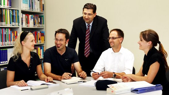 Fünf Personen sitzen an einem Tisch in einer Bibliothek und diskutieren. Ein Mann steht und leitet das Gespräch. Bücherregale im Hintergrund.