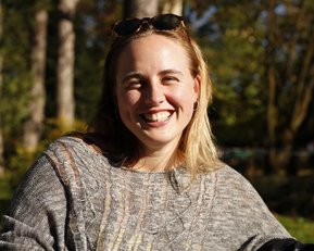 Eine lächelnde Frau mit blonden Haaren trägt einen grauen Pullover und sitzt im Freien, umgeben von Bäumen.