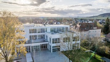 Moderne, mehrstöckige Gebäude mit großen Fenstern, umgeben von Bäumen und einem Parkplatz. Im Hintergrund sind Wohnhäuser und Hügel sichtbar.