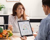 Eine Frau zeigt einem Mann auf einem Tablet eine Ernährungstabelle, während sie am Tisch mit Obst und Gemüse sitzt.
