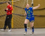 Eine Handballspielerin in blauer Uniform jubelt nach einem Tor, während eine Torhüterin in roter Kleidung im Hintergrund steht. Die Szene spielt sich in einer Sporthalle ab.