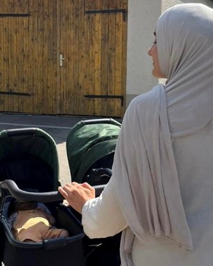 Eine Frau in einem Kopftuch schiebt einen Kinderwagen auf einer Straße vor einem Haus mit Holzgarage und blauem Himmel.