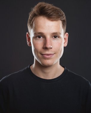 Ein junger Mann mit kurzem, braunem Haar und einem neutralen Gesichtsausdruck trägt ein schwarzes T-Shirt. Der Hintergrund ist dunkel und hebt seine Gesichtszüge hervor.