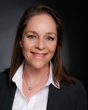 Porträt einer Frau mit langen, braunen Haaren, die ein weißes Hemd und einen schwarzen Blazer trägt. Sie lächelt freundlich in die Kamera.