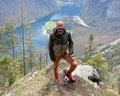 Eine Person steht auf einem Felsen mit Blick auf einen malerischen See und umliegende Berge. Die Landschaft ist von Bäumen umgeben, und der Himmel ist teilweise bewölkt.