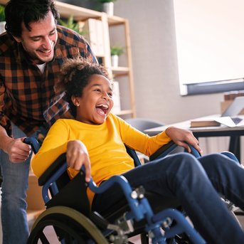 Heilpädagogen arbeiten eng mit Menschen zusammen, deren Entwicklung besondere Aufmerksamkeit und Unterstützung erfordert. Ein Mann schiebt ein Kind im Rollstuhl, während das Kind fröhlich lacht. Beide befinden sich in einem modernen Raum mit Möbeln und Pflanzen im Hintergrund.