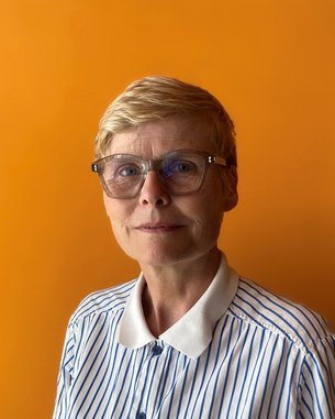 Eine Frau mit kurzen blonden Haaren und Brille trägt ein gestreiftes Hemd vor einem orangefarbenen Hintergrund.