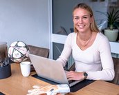 Eine Frau sitzt an einem Tisch mit einem Laptop, einem Fußball und einer Tasse. Sie trägt einen hellen Pullover und hat Handschuhe für Torhüter neben sich.