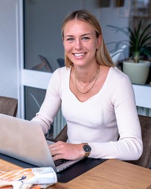 Eine Frau sitzt an einem Tisch mit einem Laptop, einem Fußball und einer Tasse. Sie trägt einen hellen Pullover und hat Handschuhe für Torhüter neben sich.