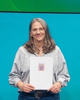 Eine Frau hält eine Urkunde in der Hand und steht vor einem Bildschirm mit dem Text "formnext". Sie trägt ein graues Oberteil und Jeans.