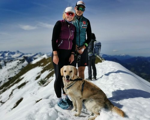 Zwei Personen stehen auf einem schneebedeckten Gipfel, umgeben von Bergen. Eine Person trägt eine Sonnenbrille und eine Weste, die andere einen Sportanzug. Ein Labrador sitzt vor ihnen.