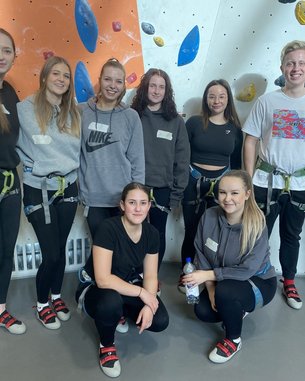 Eine Gruppe von acht Personen in Kletterausrüstung steht vor einer Kletterwand. Alle tragen Kletterschuhe und Gurte.