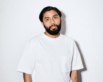 Ein Mann mit Bart trägt ein weißes T-Shirt und schwarze Hose. Er steht vor einem neutralen Hintergrund, die Hände lässig in den Hosentaschen.