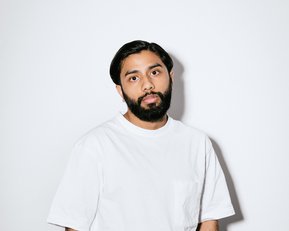 Ein Mann mit Bart trägt ein weißes T-Shirt und schwarze Hose. Er steht vor einem neutralen Hintergrund, die Hände lässig in den Hosentaschen.