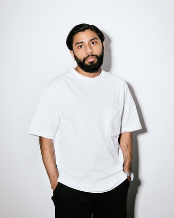 Ein Mann mit Bart trägt ein weißes T-Shirt und schwarze Hose. Er steht vor einem neutralen Hintergrund, die Hände lässig in den Hosentaschen.