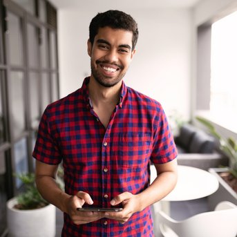 Junger Mann mit dunklen Haaren trägt ein kariertes Hemd und hält ein Smartphone in der Hand, steht in einem modernen Raum mit Pflanzen.
