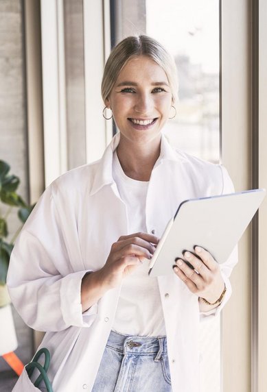 Eine lächelnde Frau in einem weißen Laborkittel hält ein Tablet in der Hand, während sie an einem Fenster steht. Im Hintergrund ist eine Pflanze und ein urbaner Ausblick zu sehen.