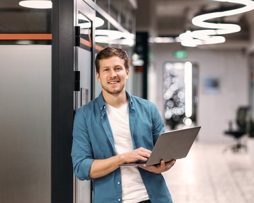 Ein Mann steht in einem modernen Büroflur und hält einen Laptop in der Hand. Er trägt ein hellblaues Hemd und lächelt.