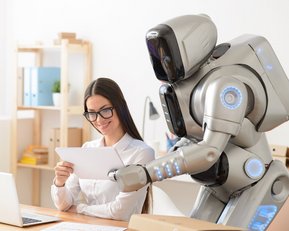 Eine Frau mit Brille sitzt an einem Schreibtisch und betrachtet ein Blatt Papier, während ein humanoider Roboter ihr gegenübersteht und das Blatt hält. Ein Laptop ist ebenfalls auf dem Tisch.