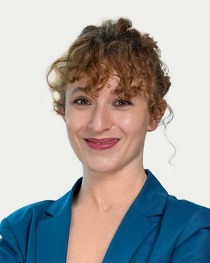 Eine lächelnde Frau mit lockigem Haar trägt einen blauen Blazer und steht mit verschränkten Armen vor einem hellen Hintergrund.
