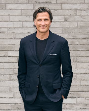 Mann in einem dunklen Anzug steht mit verschränkten Händen vor einer grauen Ziegelwand.