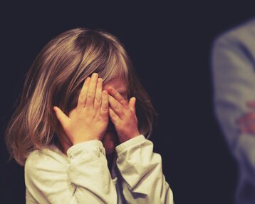 Ein kleines Mädchen mit schulterlangen Haaren hält sich die Hände vor das Gesicht, während zwei Personen im Hintergrund stehen.