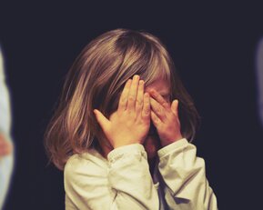 Ein kleines Mädchen mit schulterlangen Haaren hält sich die Hände vor das Gesicht, während zwei Personen im Hintergrund stehen.