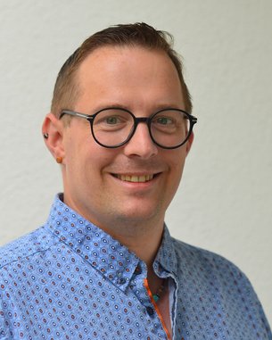 Ein lächelnder Mann mit Brille trägt ein gemustertes Hemd. Er hat kurze, hellbraune Haare und Ohrstecker. Der Hintergrund ist neutral und hell.